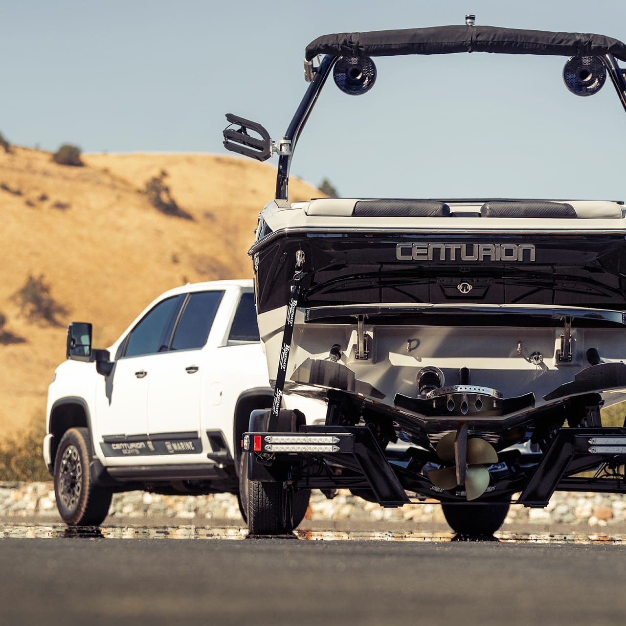 A white pickup truck is parked next to a Centurion boat on a trailer with a dry, hilly landscape in the background.