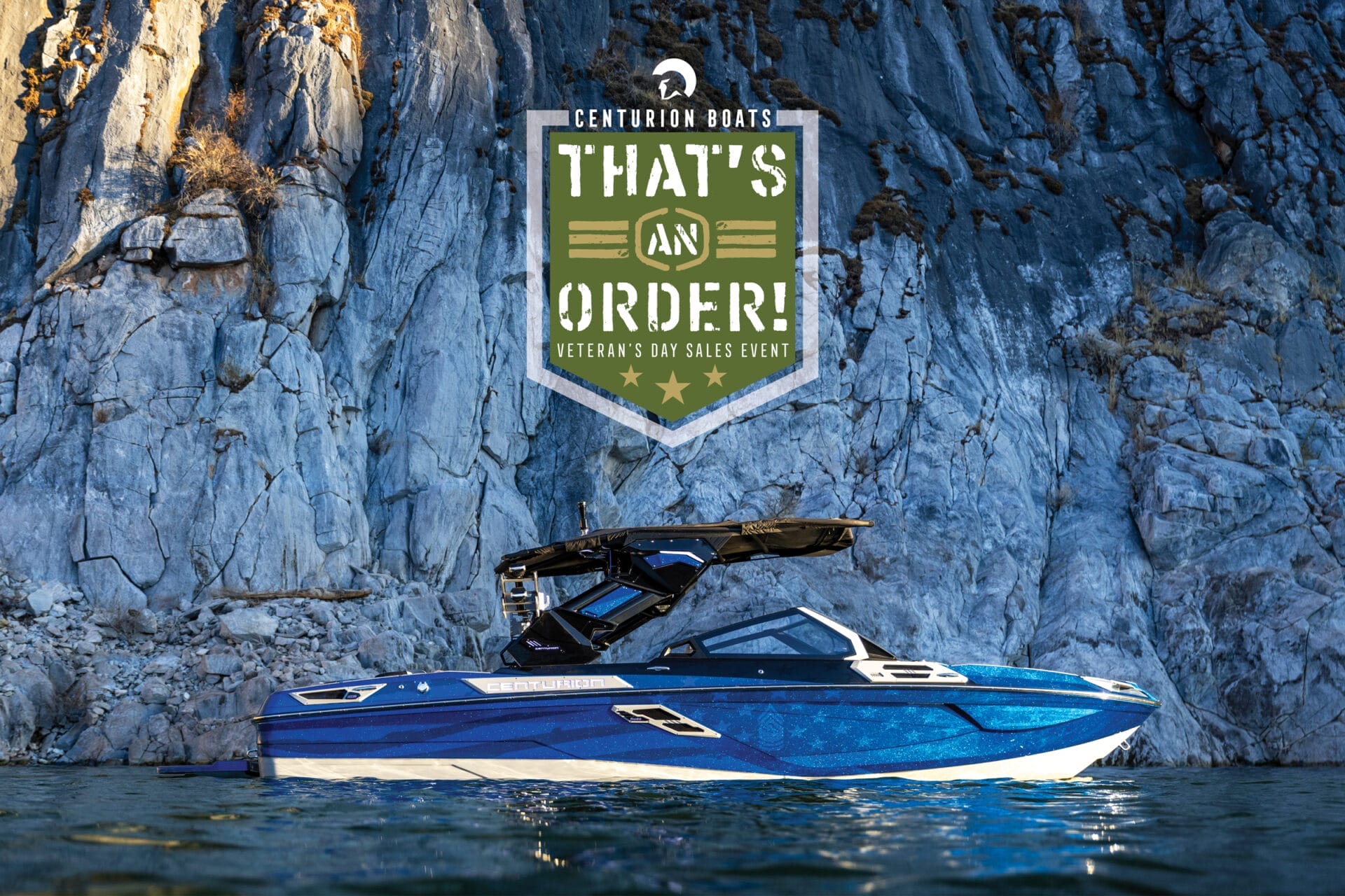 A blue and white boat floats on water near a rocky cliff. A large green and white sign above reads "Centurion Boats: That's an Order! Veteran’s Day Sales Event.