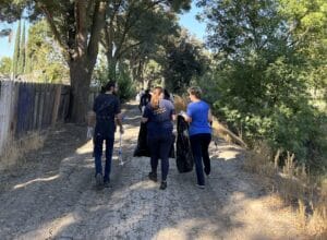 A group of people walk down a tree-lined dirt path, carrying trash bags, likely participating in a cleanup effort.