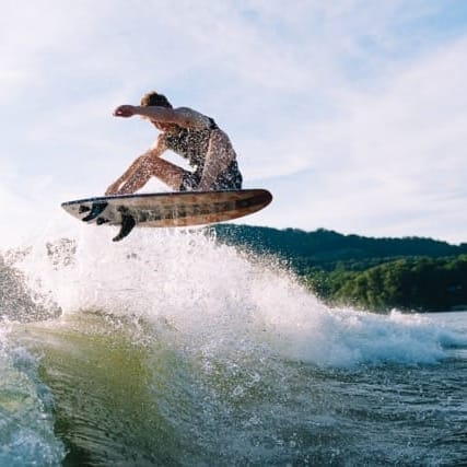 A person on a surfboard catches air off a wave, with a distant shoreline and a hilly backdrop under a partly cloudy sky.