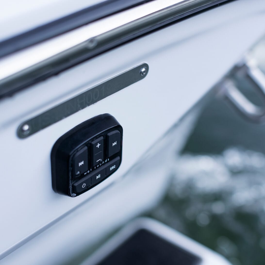 Close-up of a boat's control panel with buttons for play, pause, volume, and track navigation, mounted on a white surface with water visible in the background.