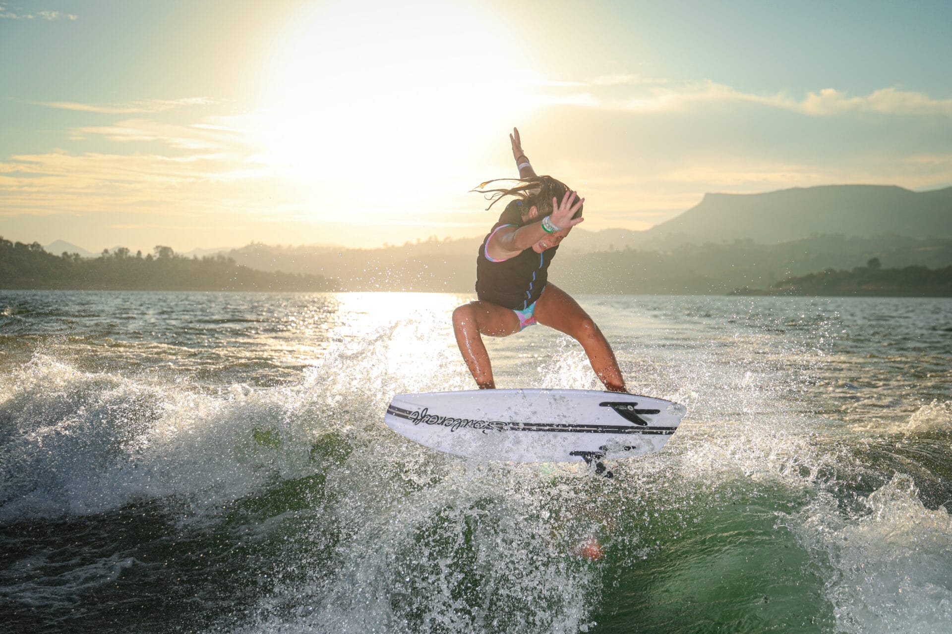 Person surfing on a wave during sunset, with water splashing around and mountains in the background.