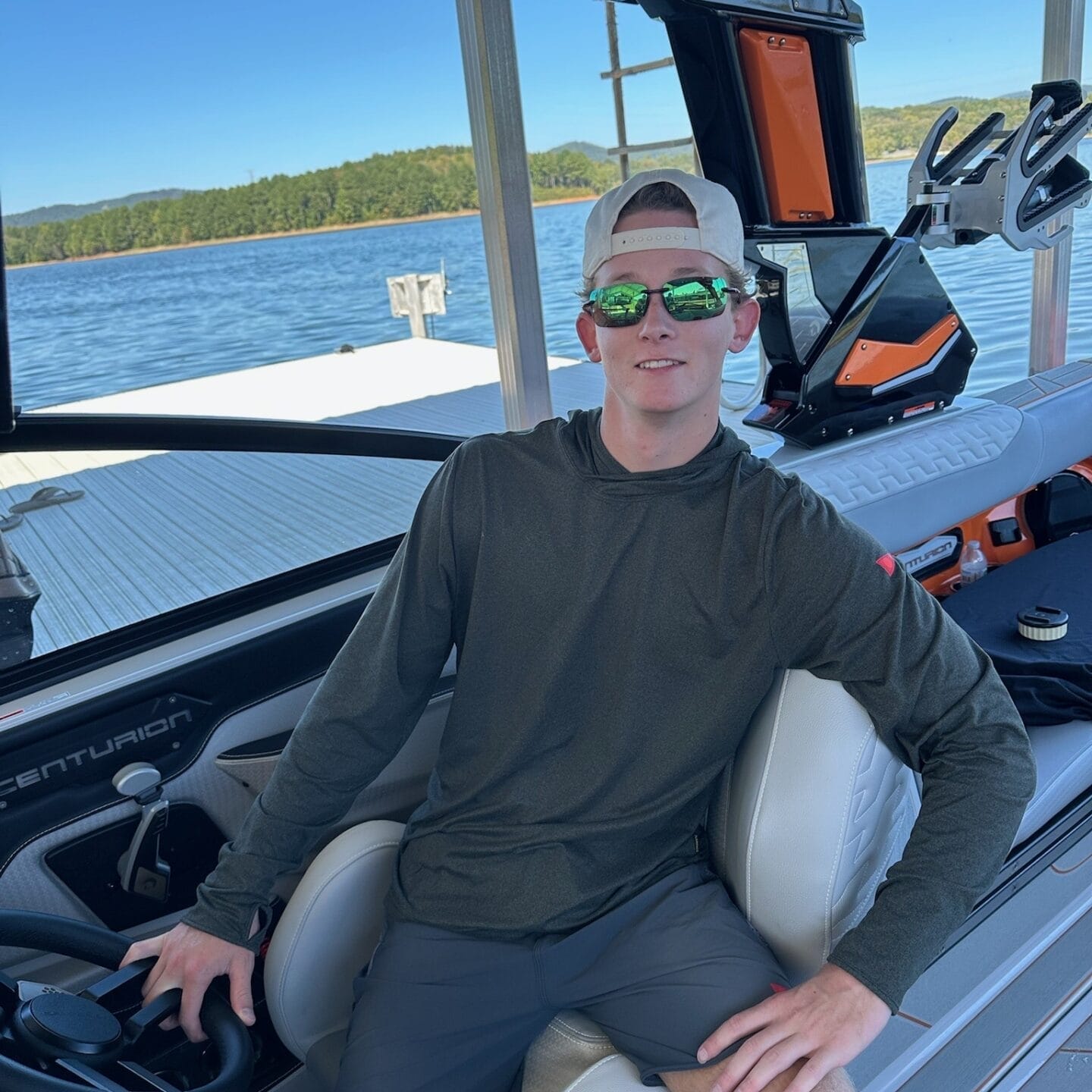 Young man wearing sunglasses and a hat sits at the helm of a boat on a lake with trees in the background.