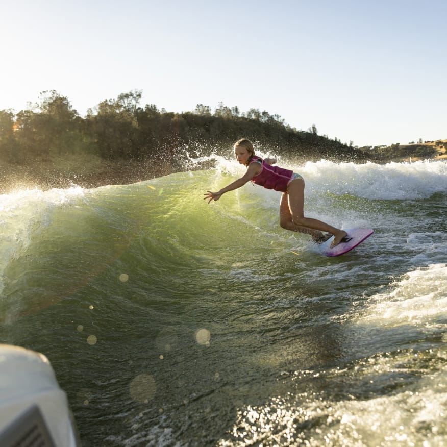 A person in a life jacket expertly surfs on a wave behind a sleek Centurion Fe Series boat on a sunny day, with trees and hills providing a picturesque backdrop.