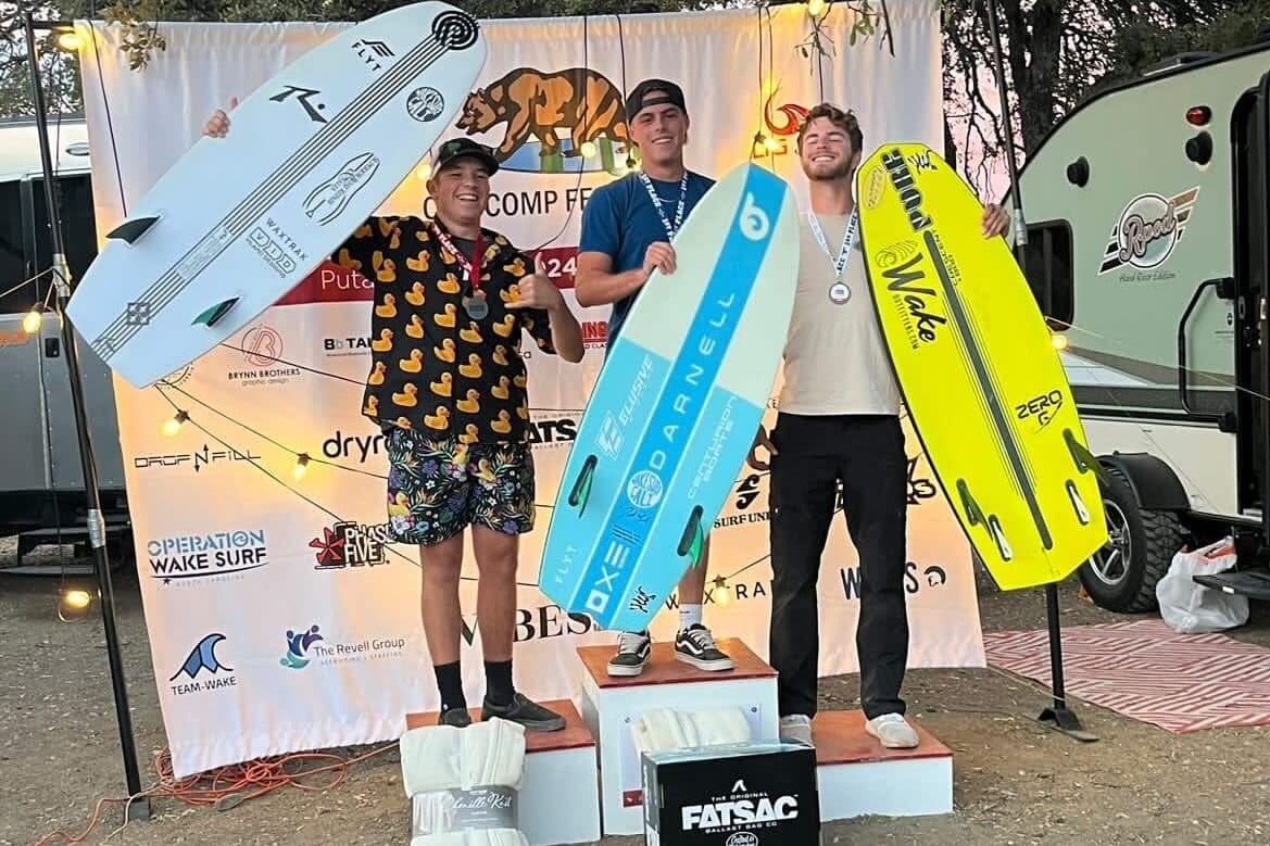 Three people stand on podiums holding large wakeboards in front of a branded backdrop at a competition event.