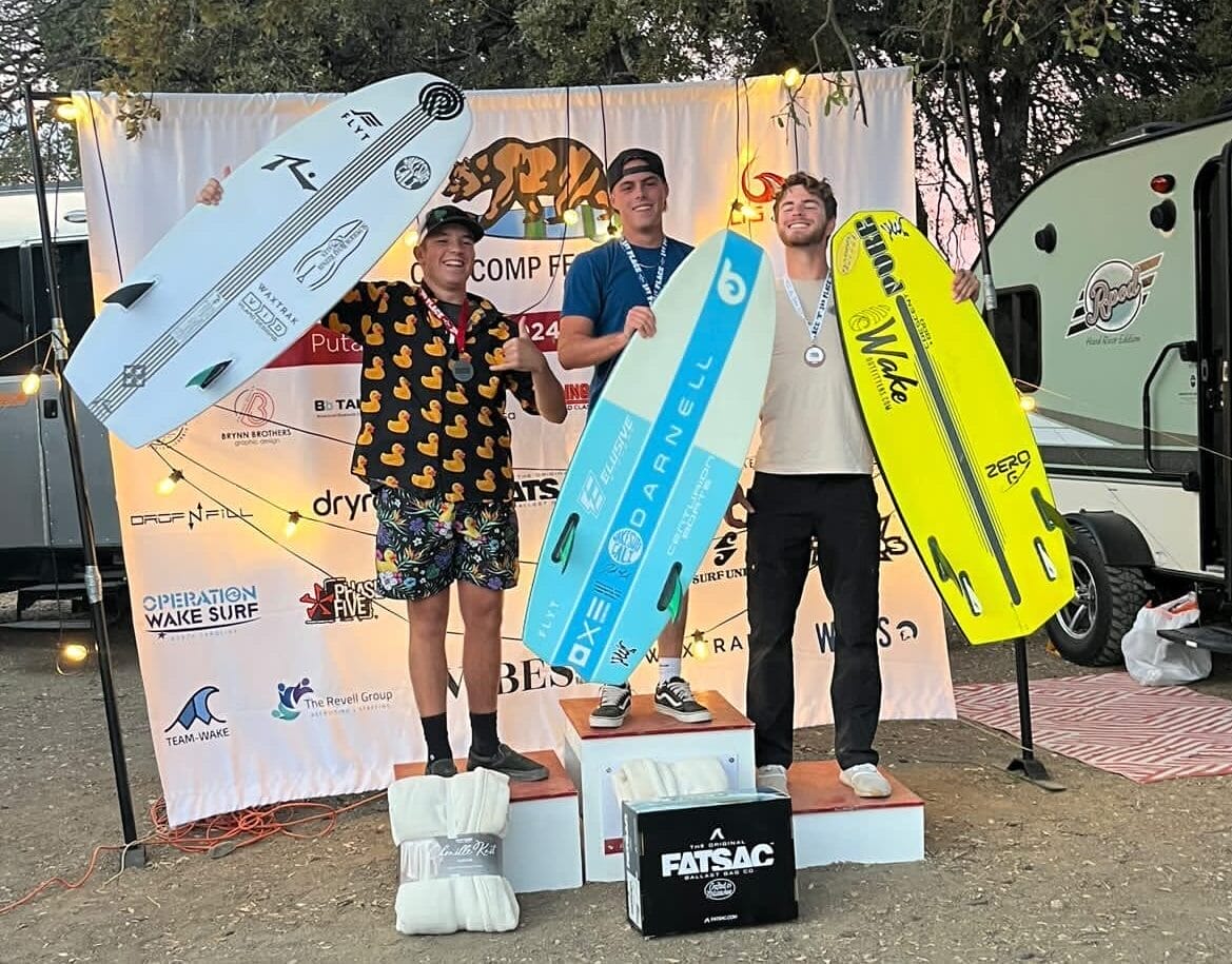 Three people stand on podiums holding large wakeboards in front of a branded backdrop at a competition event.