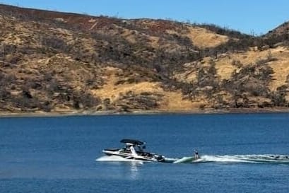 A person water skiing on a lake behind a boat on a sunny day, with dry, rocky hills in the background.