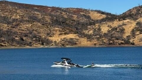 A person water skiing on a lake behind a boat on a sunny day, with dry, rocky hills in the background.