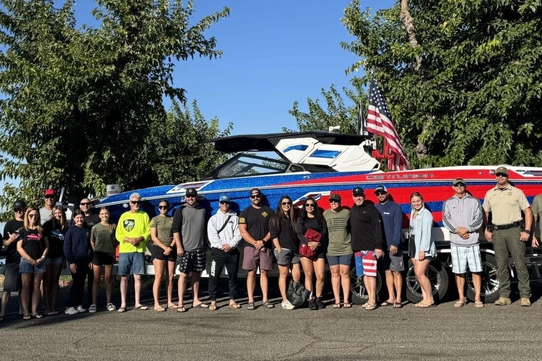 A group of people stands in front of a large blue and red boat on a trailer, with trees in the background and a clear blue sky above.