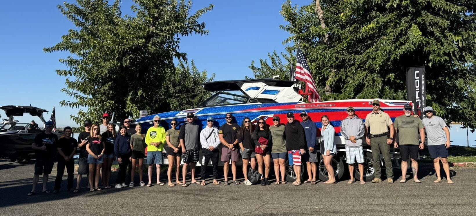 A group of people stands in front of a large blue and red boat on a trailer, with trees in the background and a clear blue sky above.