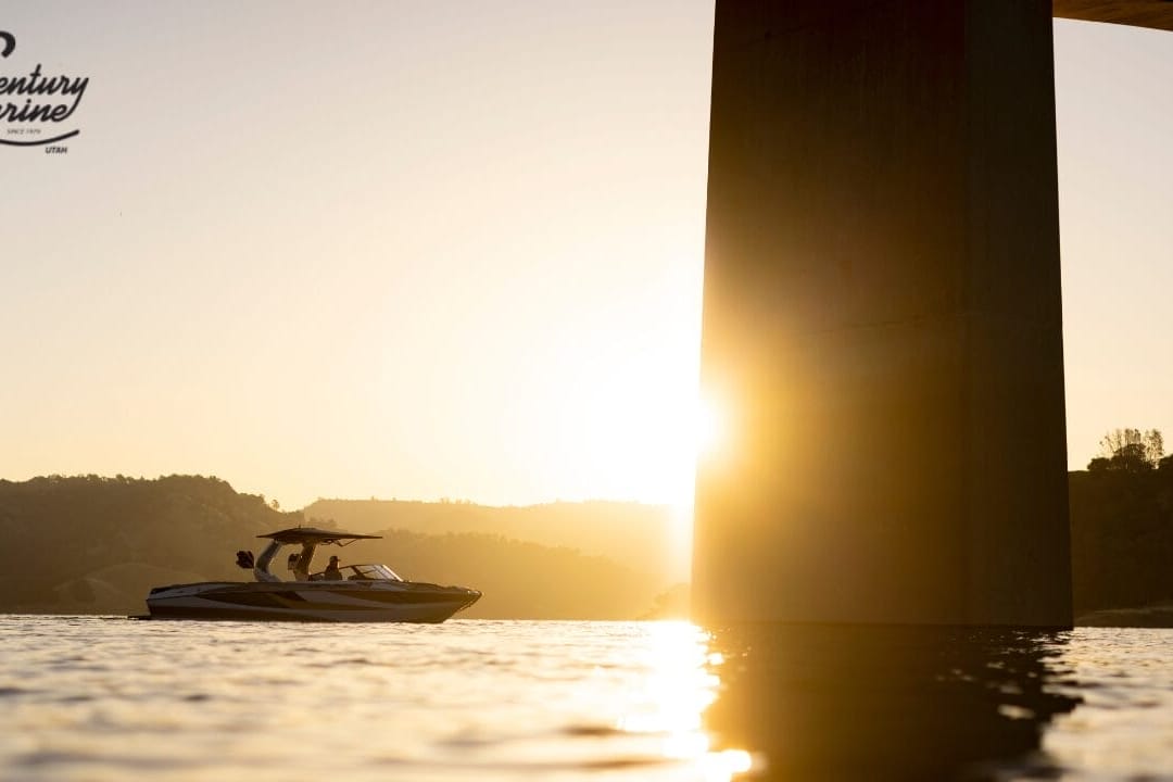 A boat cruises on a lake at sunset near a large bridge pillar, with hills in the background.