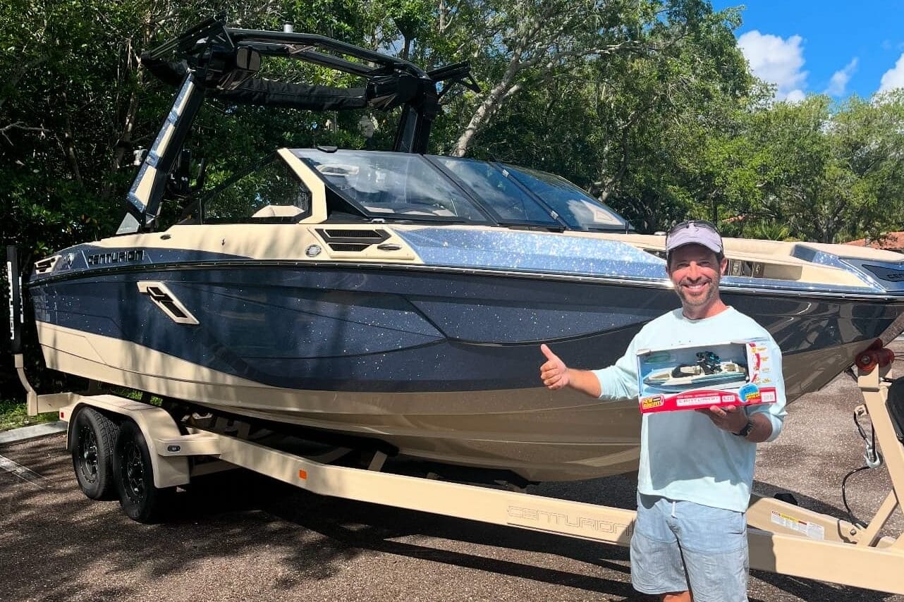 A person gives a thumbs up while holding a toy boat and standing next to a large boat on a trailer.