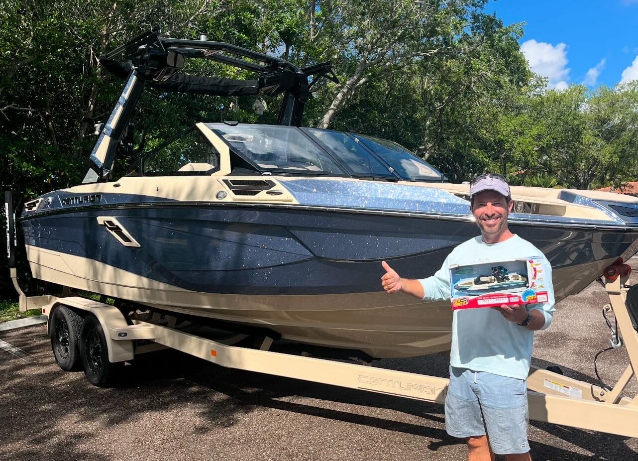 A person gives a thumbs up while holding a toy boat and standing next to a large boat on a trailer.