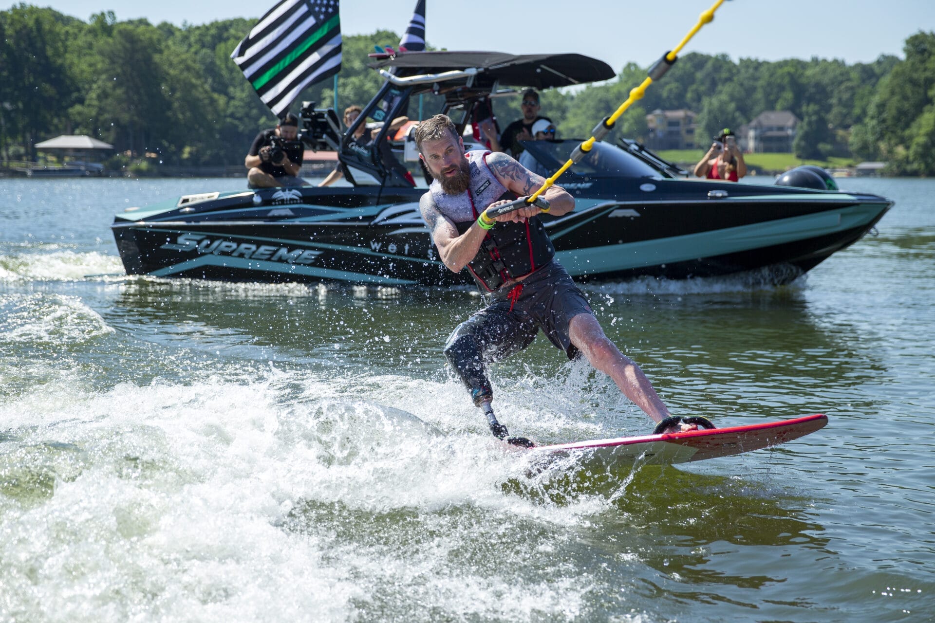 A man wakeboards on a lake, holding a rope, with a boat carrying people and flags in the background.