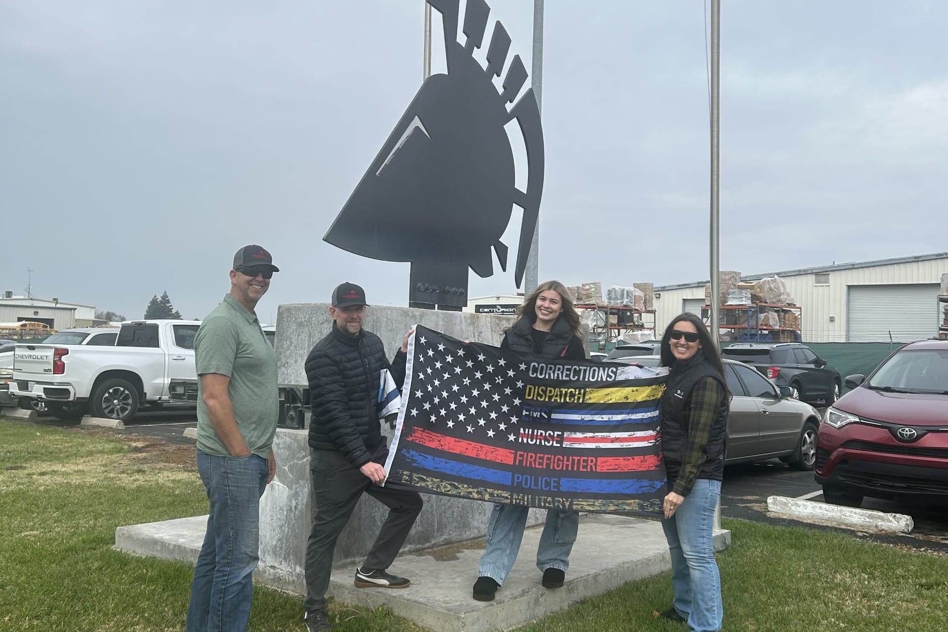 Four individuals hold a flag with various service stripes in front of a Spartan helmet statue. Vehicles and a flagpole are in the background.