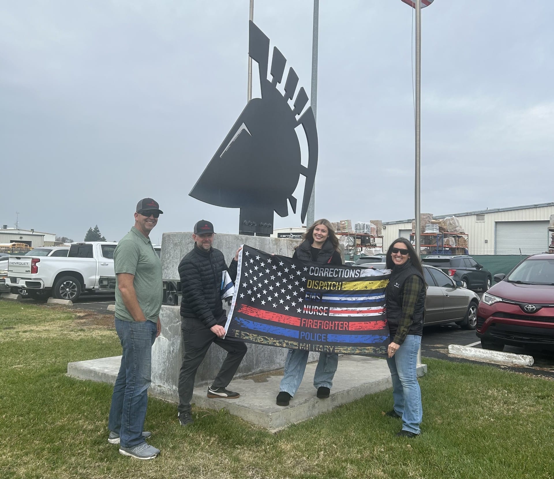 Four individuals hold a flag with various service stripes in front of a Spartan helmet statue. Vehicles and a flagpole are in the background.