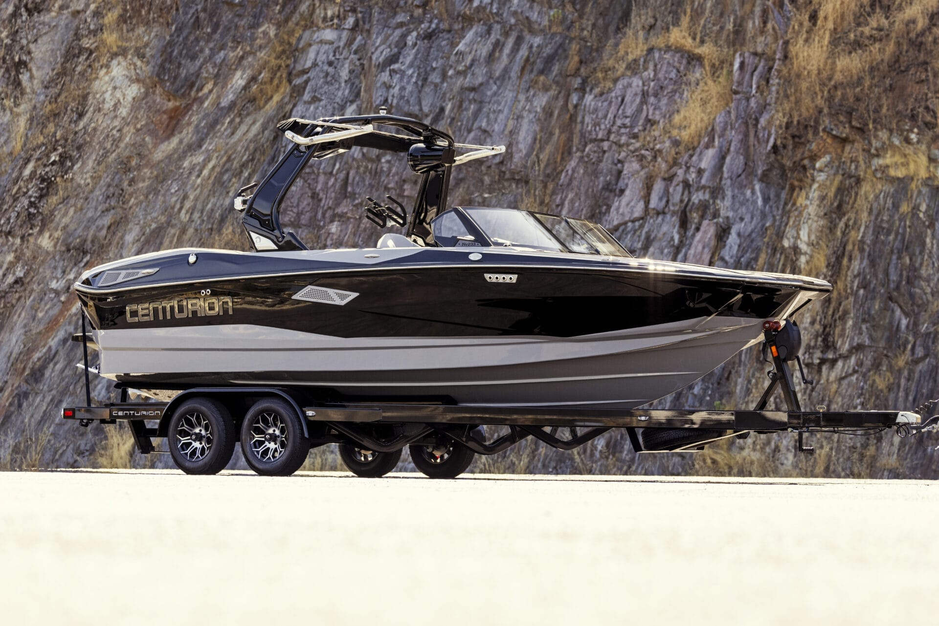 Side view of a black and white Centurion boat on a trailer, parked on a road with a rocky cliff background.