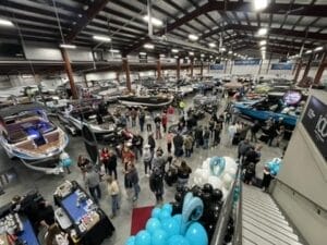 A large indoor boat show with numerous boats on display. People are gathered around, observing the boats, and there are blue and black balloons near an entrance.