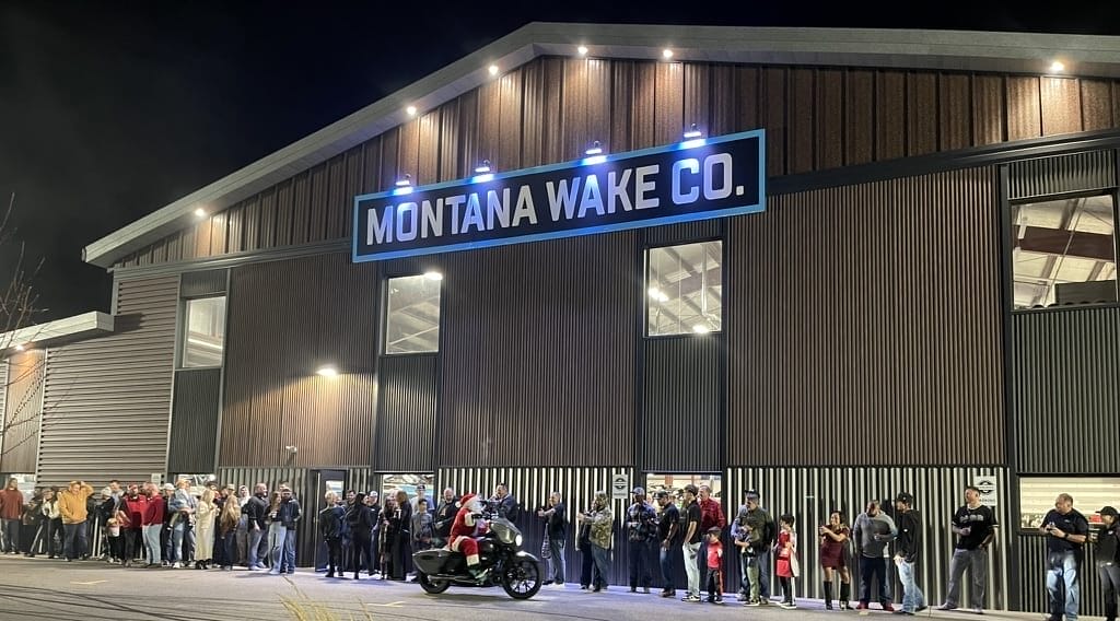 A large crowd lines up outside a building labeled "Montana Wake Co." at night, with a motorcycle in the foreground.