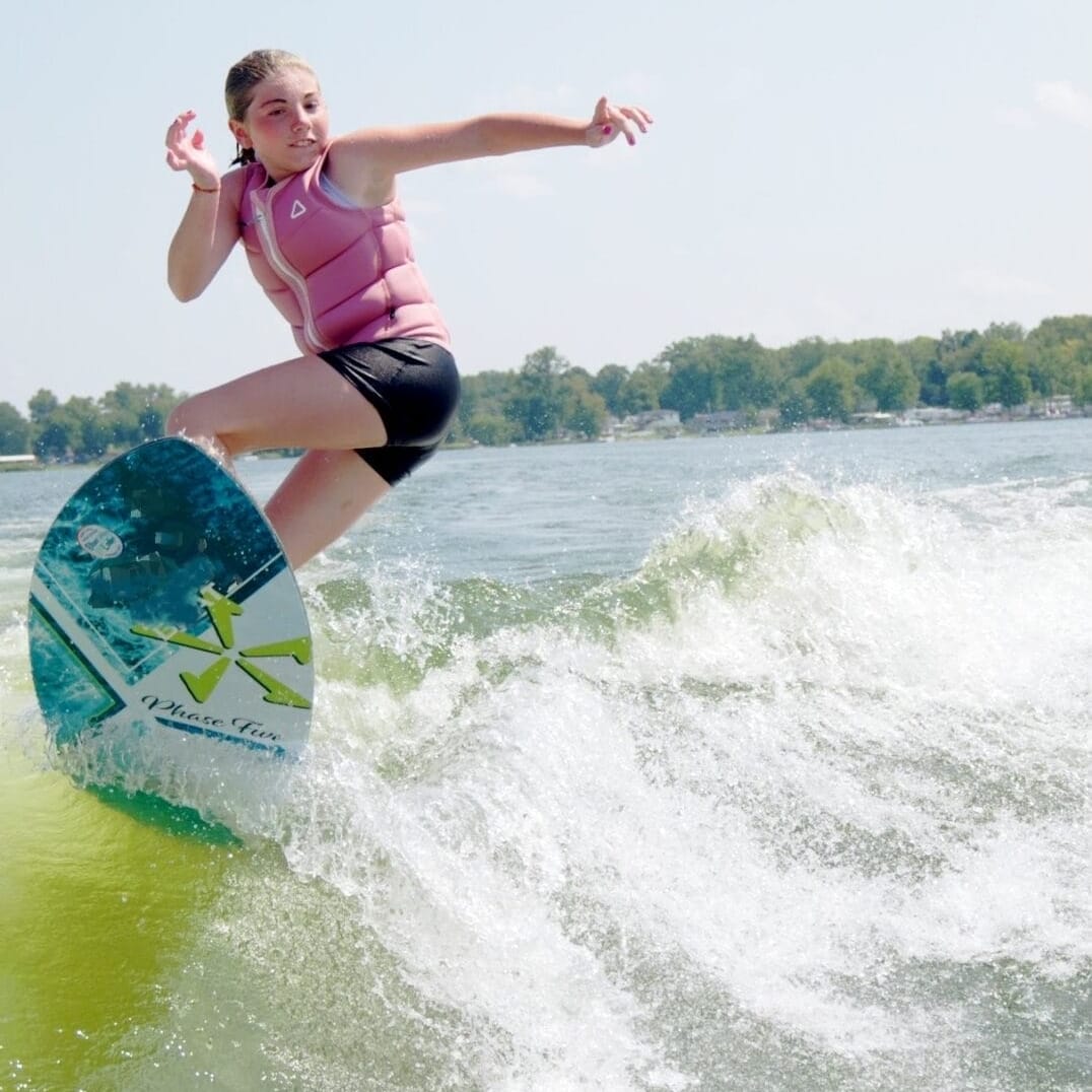 Person wakeboarding on a lake, wearing a pink life vest and navy shorts, with greenery visible in the background, against a clear sky.