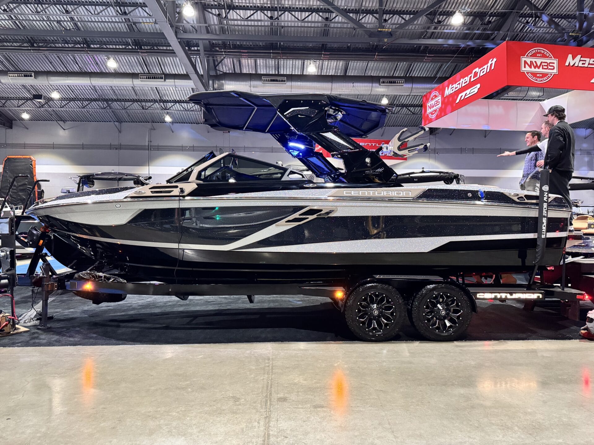 A sleek black and silver Centurion boat on display indoors, mounted on a trailer with shiny black wheels.