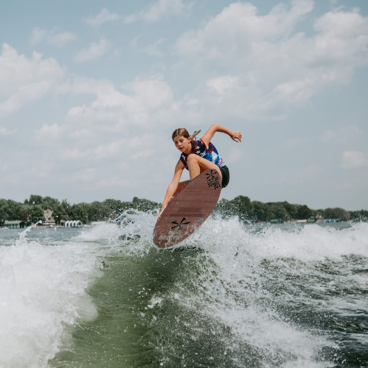 Person surfing on a wave under a partly cloudy sky, performing a jump on a red surfboard with a vibrant landscape in the background.