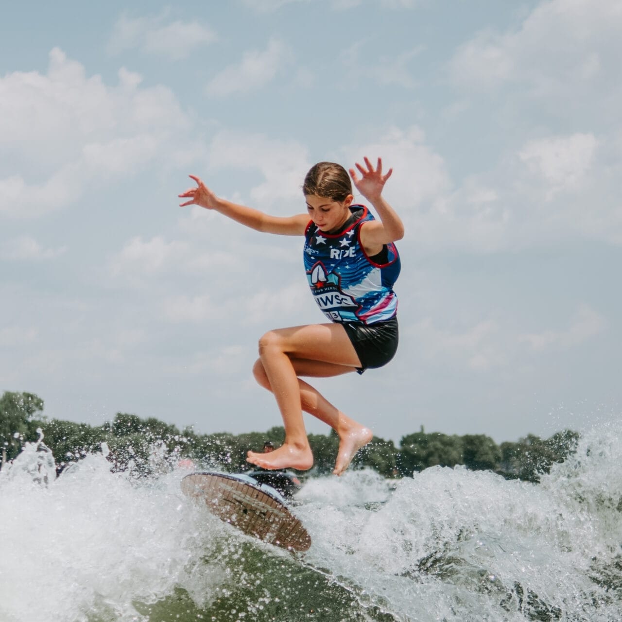 Person surfing on a wave under a blue sky with scattered clouds, wearing a colorful vest and black shorts.