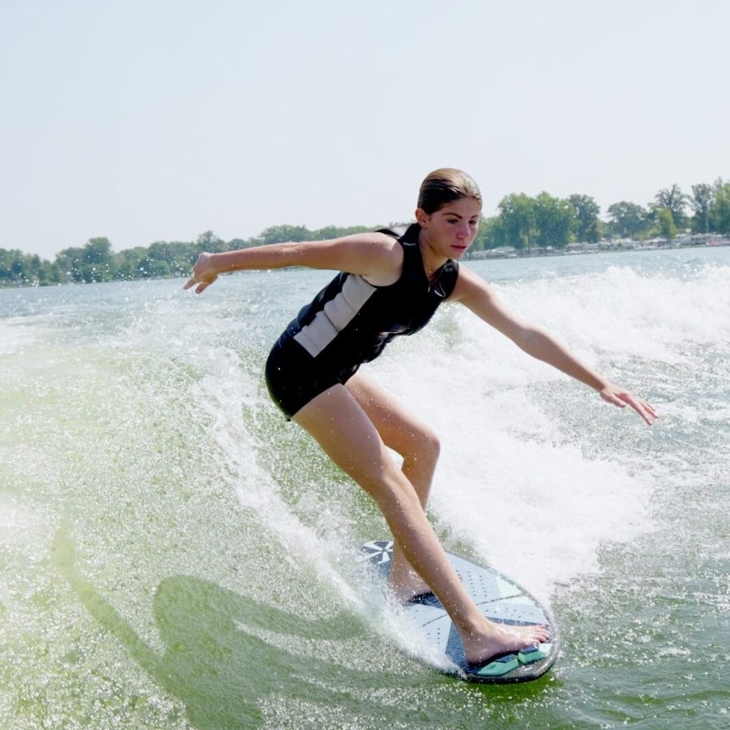 A person in a wetsuit rides a surfboard on a wave in a sunny setting, with trees visible in the background.