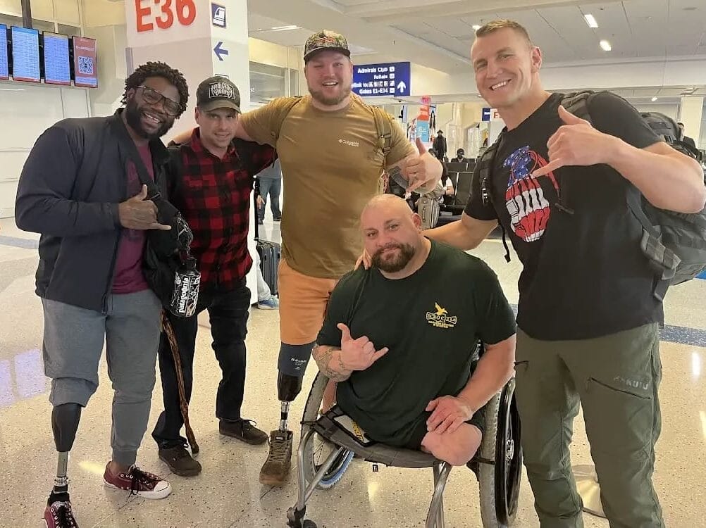 A group of five men, some with prosthetic legs, pose and smile at an airport terminal. They are casually dressed and making hand gestures. Airport signage is visible in the background.