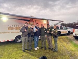 A group of six people, including uniformed officers, stand smiling in front of a Placer County Sheriff vehicle and trailer on a grassy area.