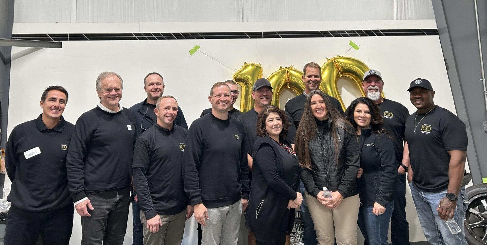 A group of thirteen people poses indoors, smiling in front of golden balloon numbers "100." They're dressed in casual and semi-formal attire in a well-lit industrial space.