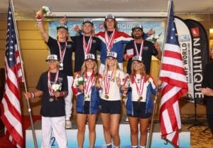 A group of athletes with medals stand on a podium. Two hold American flags. They wear matching sports attire. An official stands nearby. Event banners are in the background.