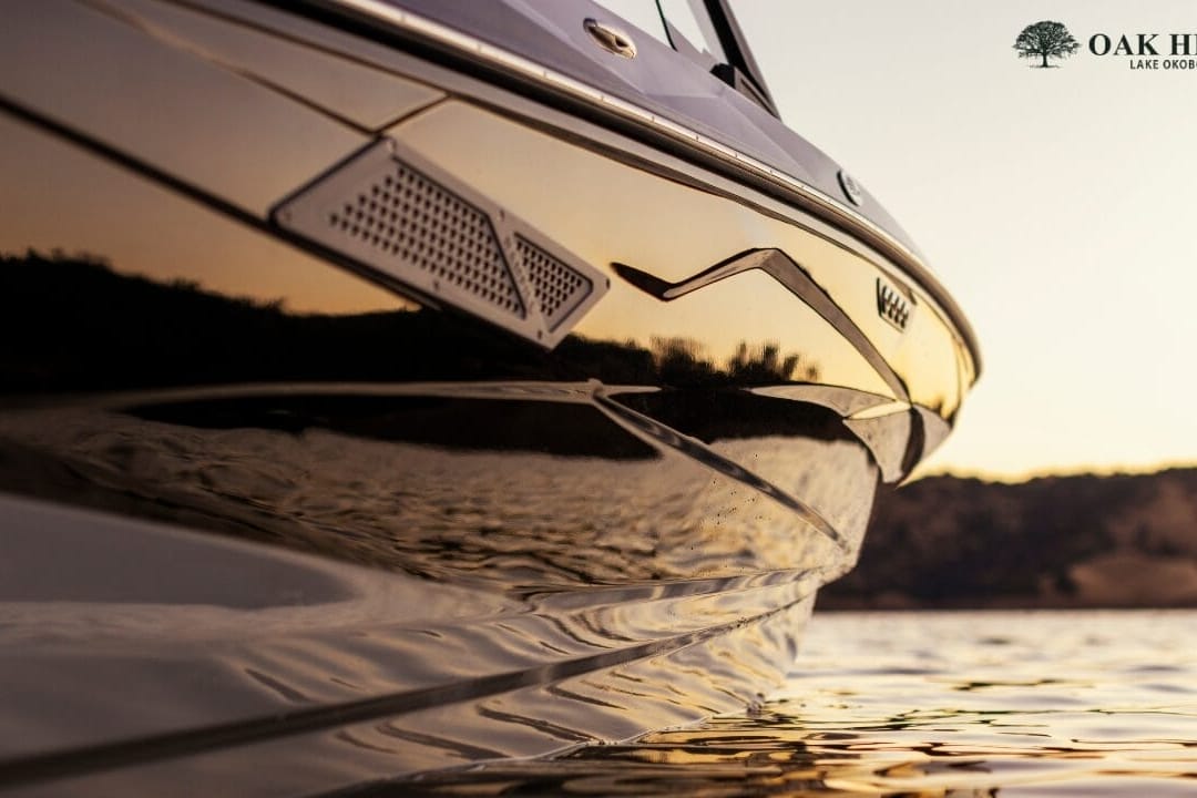 Close-up of a boat's side with reflections on the water at sunset. The Oak Hill Lake Osborn logo is in the top right corner.