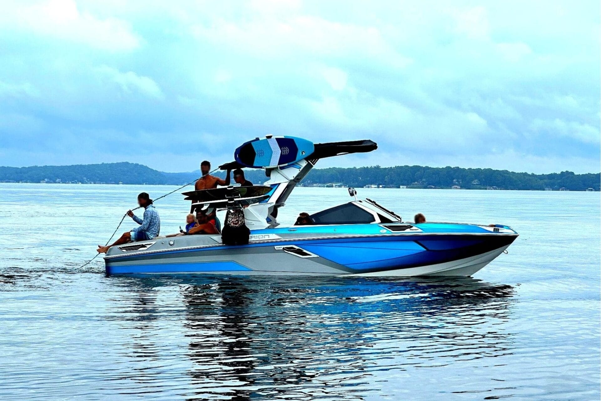 A blue and white speedboat with multiple people, including one holding a rope, is on a lake. Wakeboards are visible on the boat's rack. Trees and hills can be seen in the background under a cloudy sky.