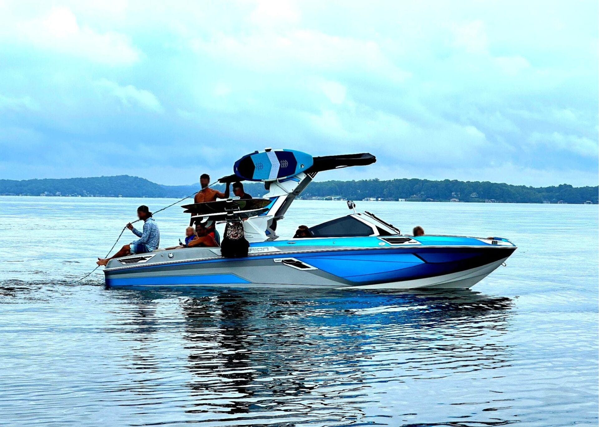 A blue and white speedboat with multiple people, including one holding a rope, is on a lake. Wakeboards are visible on the boat's rack. Trees and hills can be seen in the background under a cloudy sky.