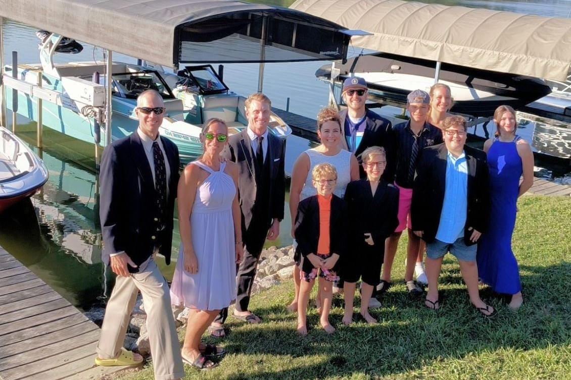 A group of people dressed in formal and casual attire pose by a dock with boats in the background.