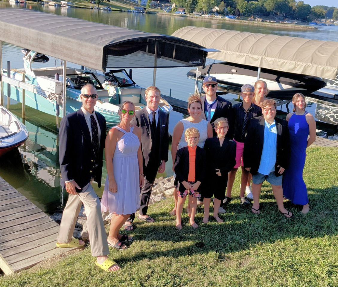 A group of people dressed in formal and casual attire pose by a dock with boats in the background.