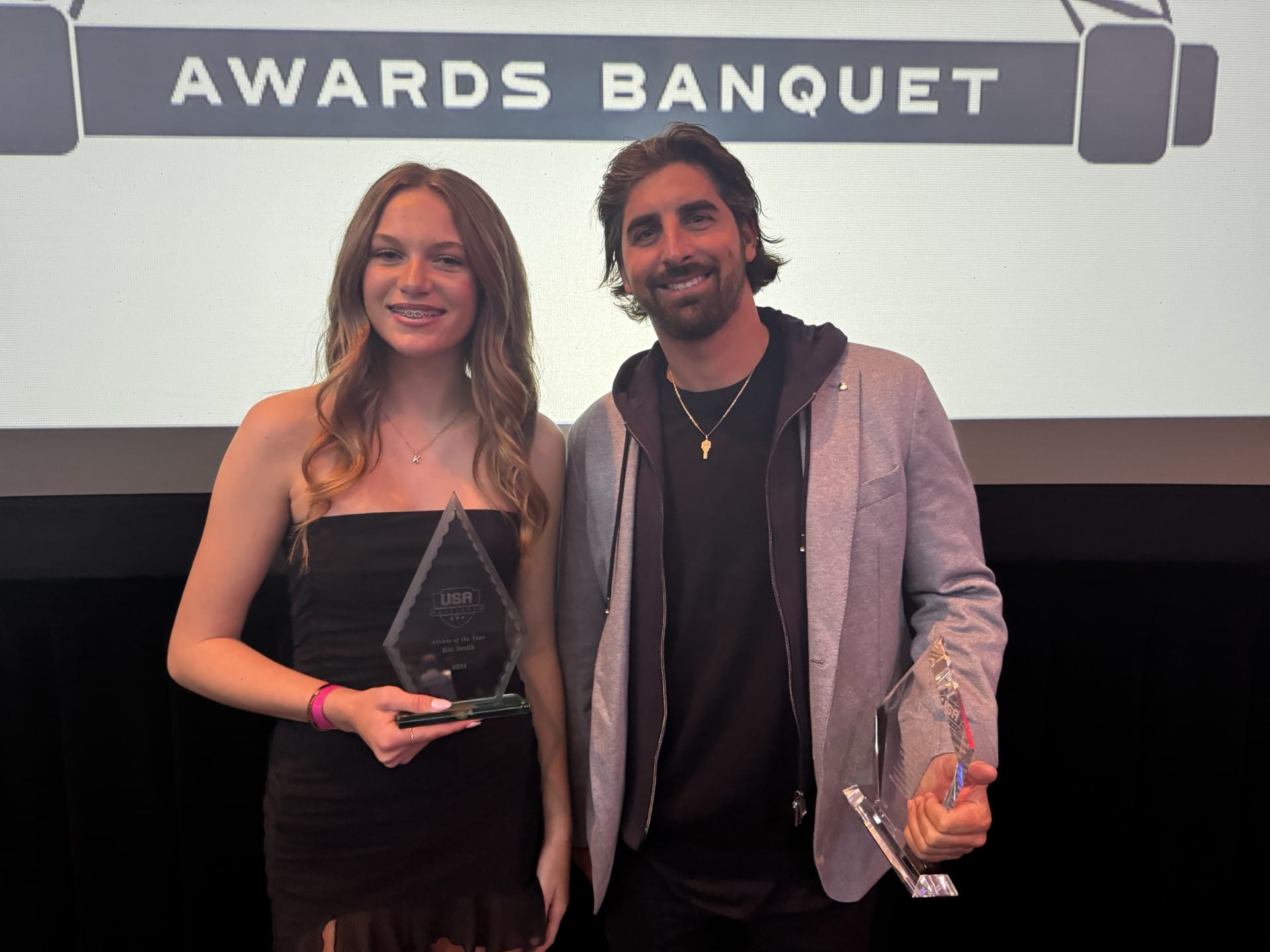 Two people holding awards and smiling at a banquet, standing in front of a backdrop that reads "Awards Banquet.