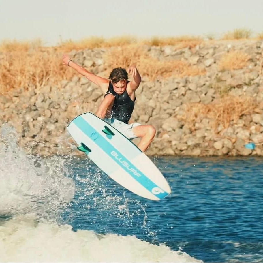 Person surfing on a blue surfboard, performing an aerial maneuver over a wave, with rocky shore and dry grass in the background.