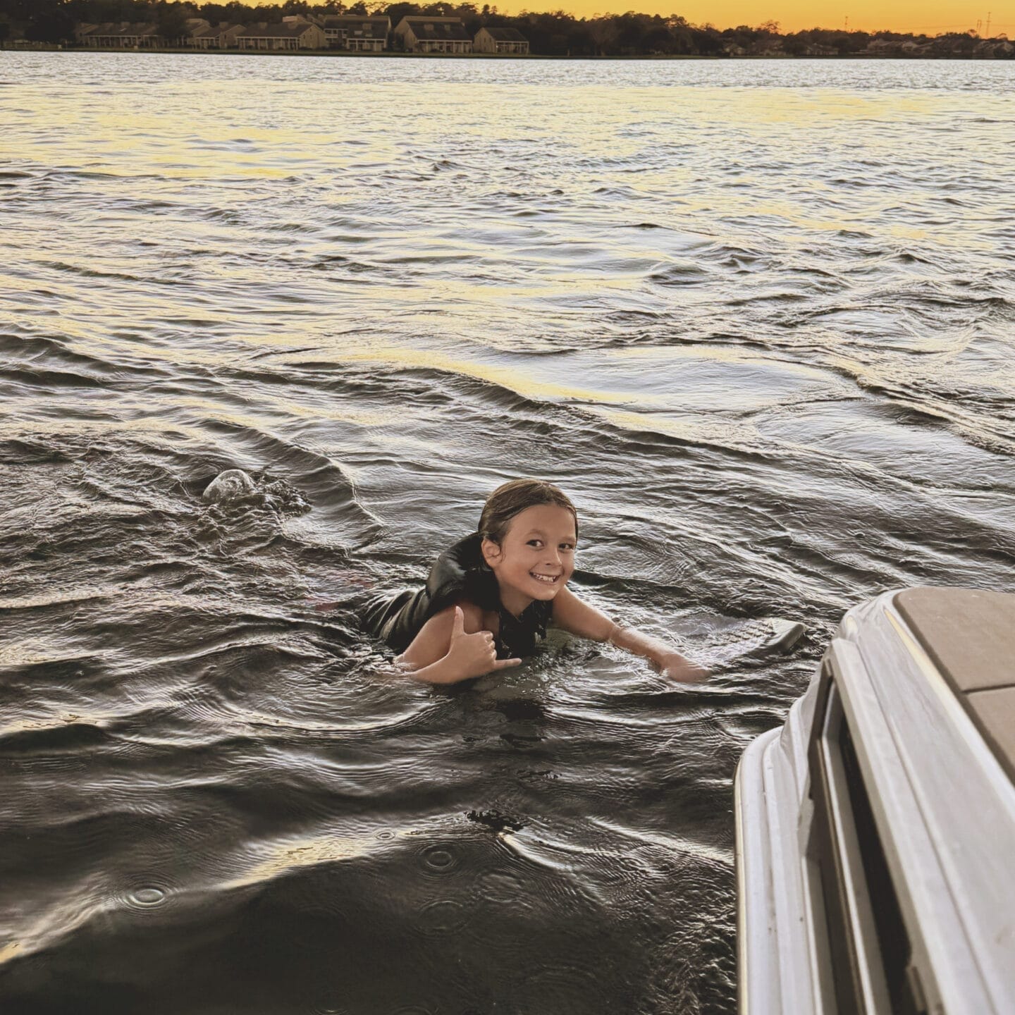 Child swimming in a lake at sunset, wearing a life vest and making a shaka sign with one hand near a boat's edge.