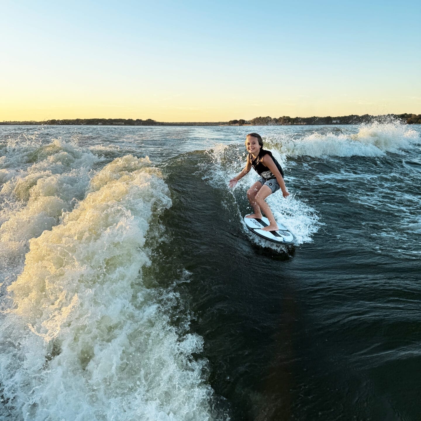 A person wakeboarding on a lake during sunset, creating waves behind a boat.