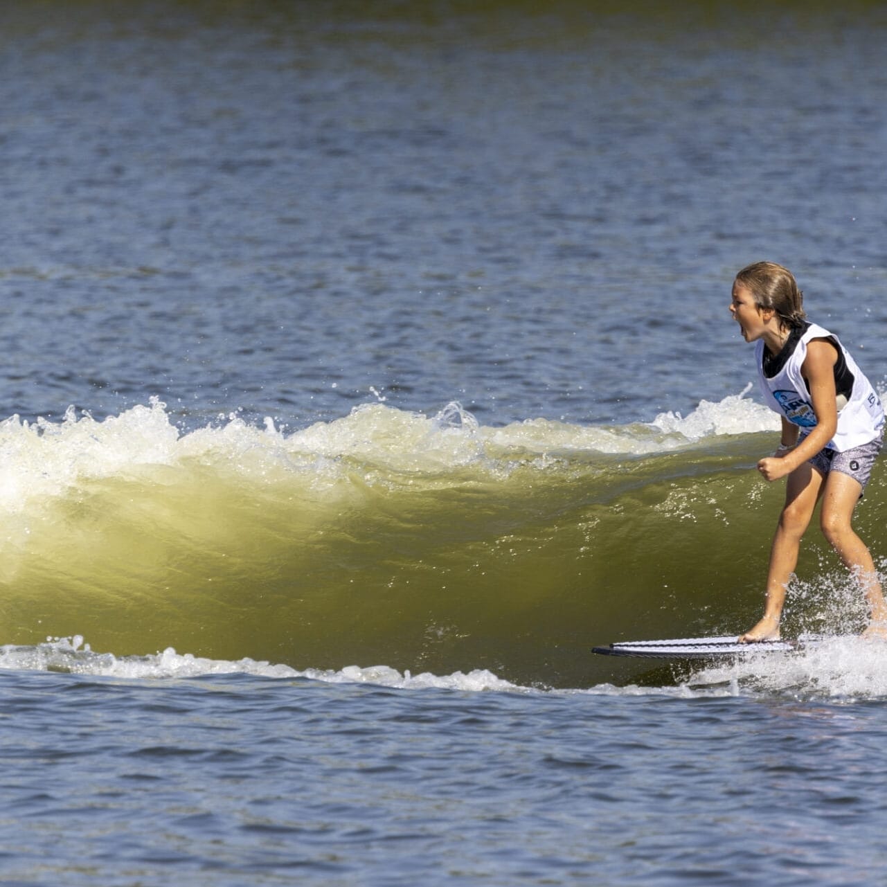 A child surfing on a wave in a body of water, wearing a life vest and shorts, with trees visible in the background.