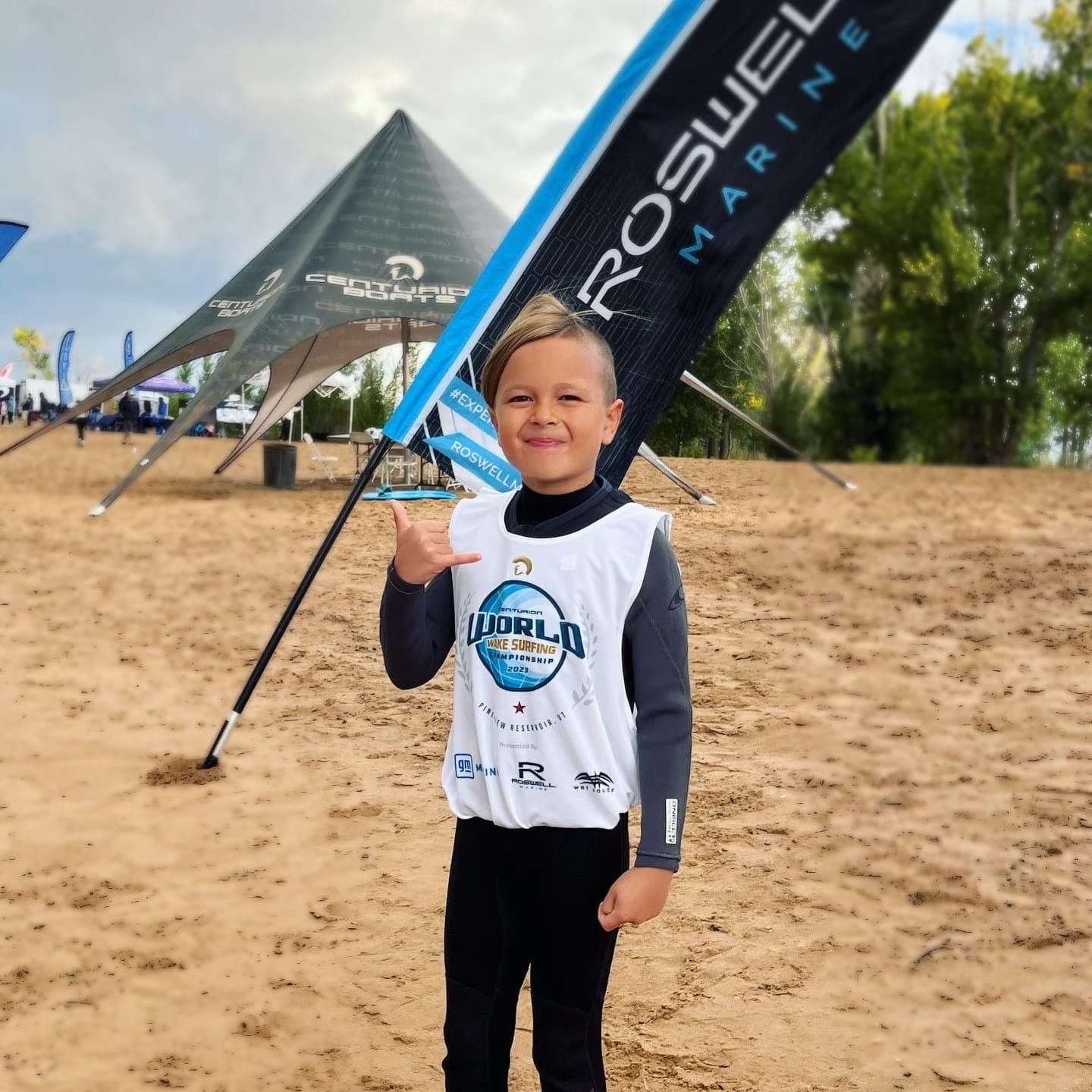 Young child in a wetsuit stands on a sandy beach, making a shaka sign. Divided banners in the background partially display 