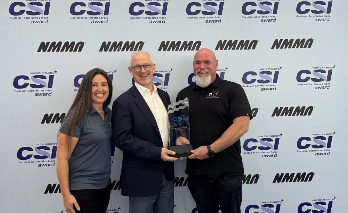 Three people stand together; the man in the center holds a trophy. They are in front of a backdrop with repeated "CSI" and "NMMA" logos.