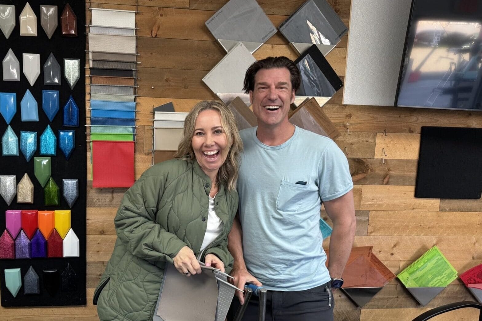 Two people smiling inside a room with samples of colorful materials displayed on the wall behind them.