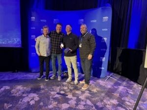 Four men stand together smiling in front of a blue backdrop with "WSIA Summit" printed on it.