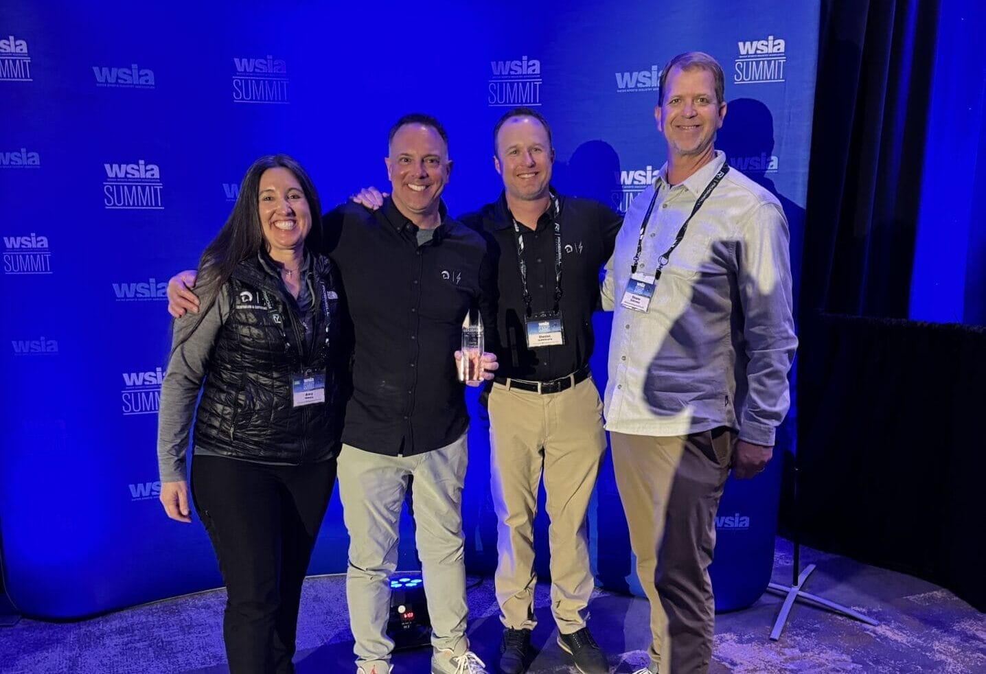 Four people smiling and posing together in front of a blue backdrop with "WSLA Summit" logos.