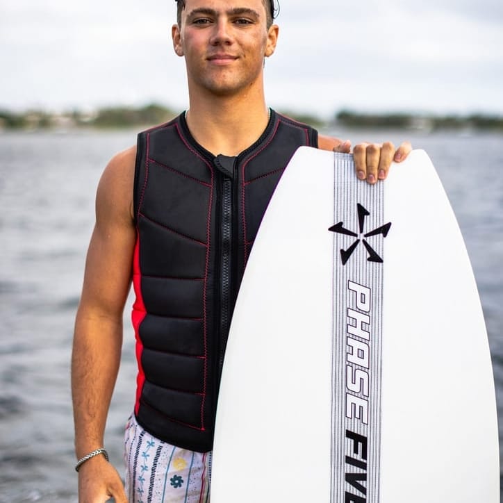 Sammy Goncalves stands by the water, holding a white surfboard vertically. Wearing a black protective vest and patterned shorts, they gaze at the overcast sky, ready for the next wave.