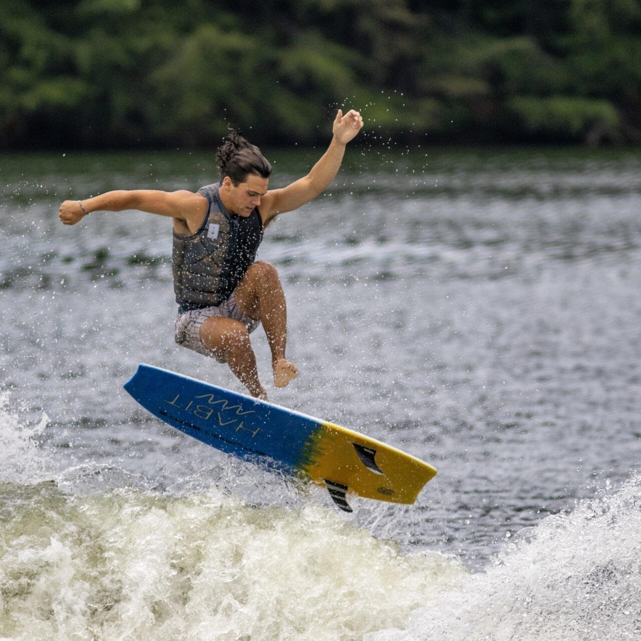 A person performs an aerial trick while wakeboarding on a body of water, surrounded by trees.