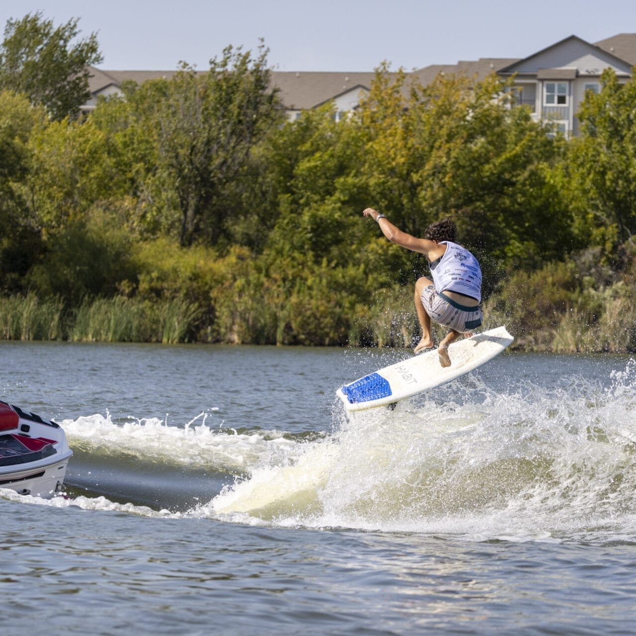 A person is wakeboarding on a lake, performing a jump behind a boat. Trees and buildings are visible in the background.